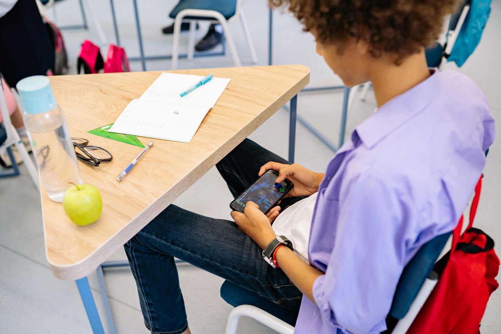 A student holding a smartphone below a school desk beside a backpack